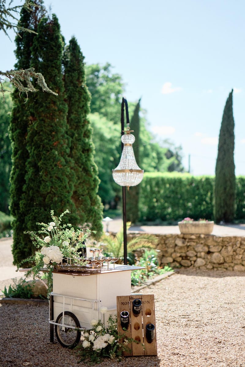 White bar cart with ranunculus, crystal chandelier on stand, and wine crate in cypress-lined garden