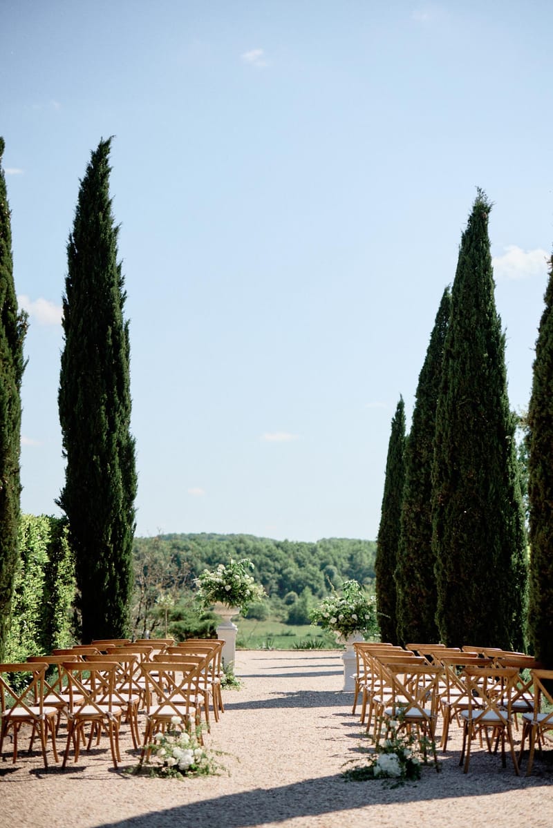 Ceremony setup on cypress-lined gravel allee with cross-back chairs and white floral aisle clusters on stone pedestals
