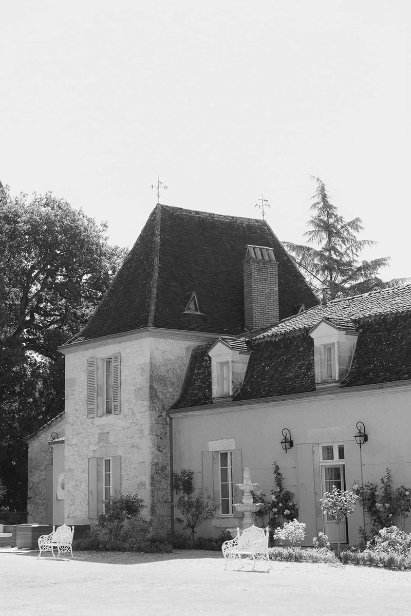 Black-and-white exterior of French chateau with corner tower, garden furniture, and stone fountain