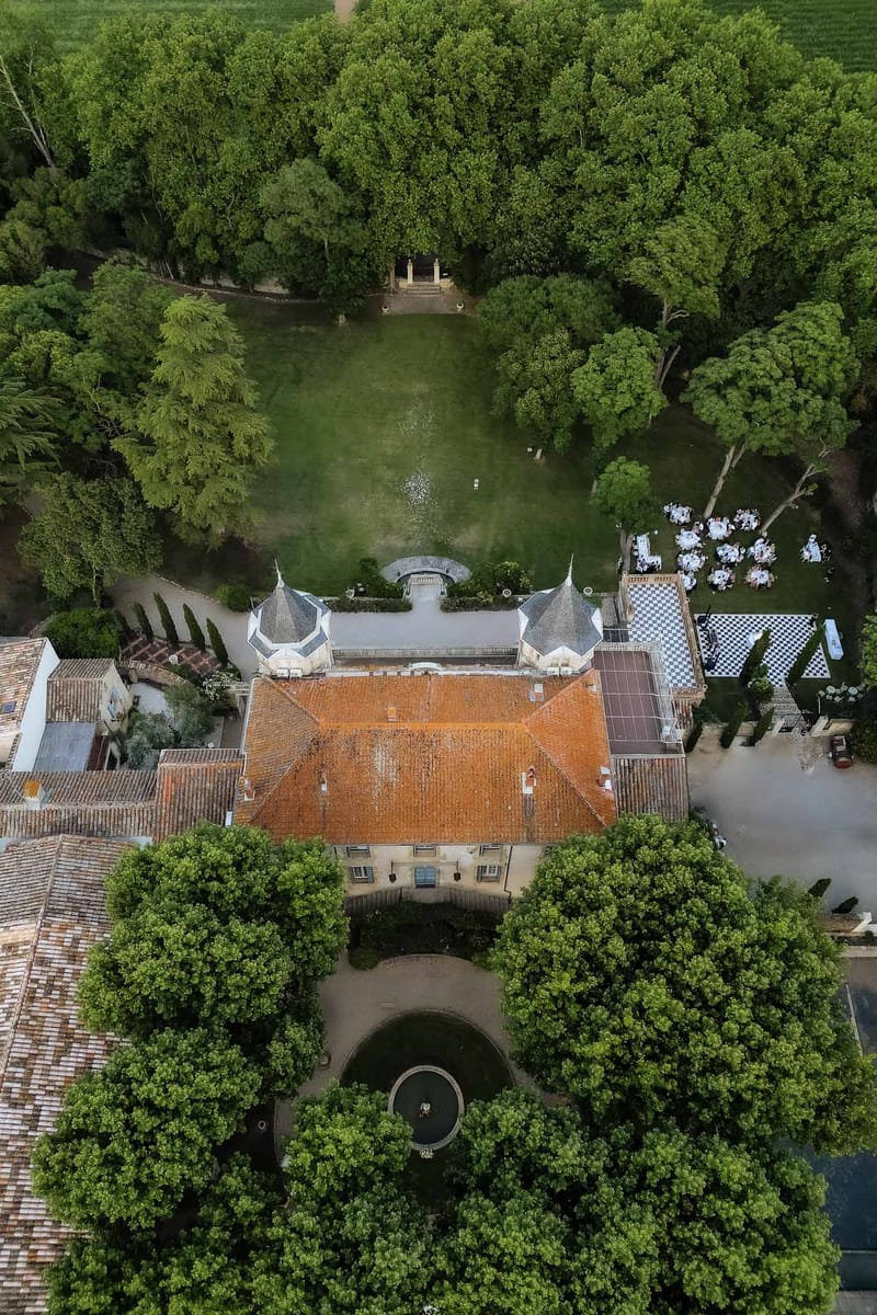 Aerial drone view of a French chateau estate with turret roofs, round reception tables, and a formal garden allee