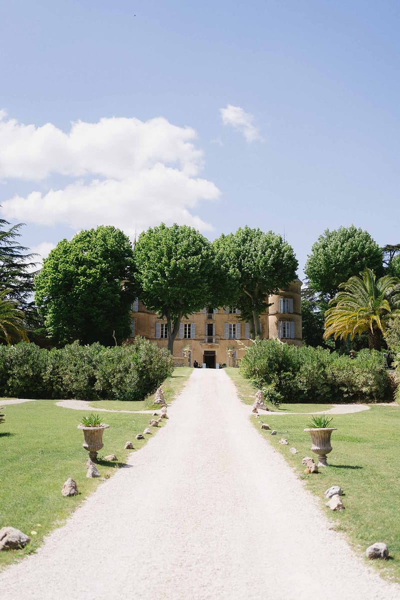 Symmetrical gravel driveway leading to golden limestone chateau with blue shutters and rounded corner tower
