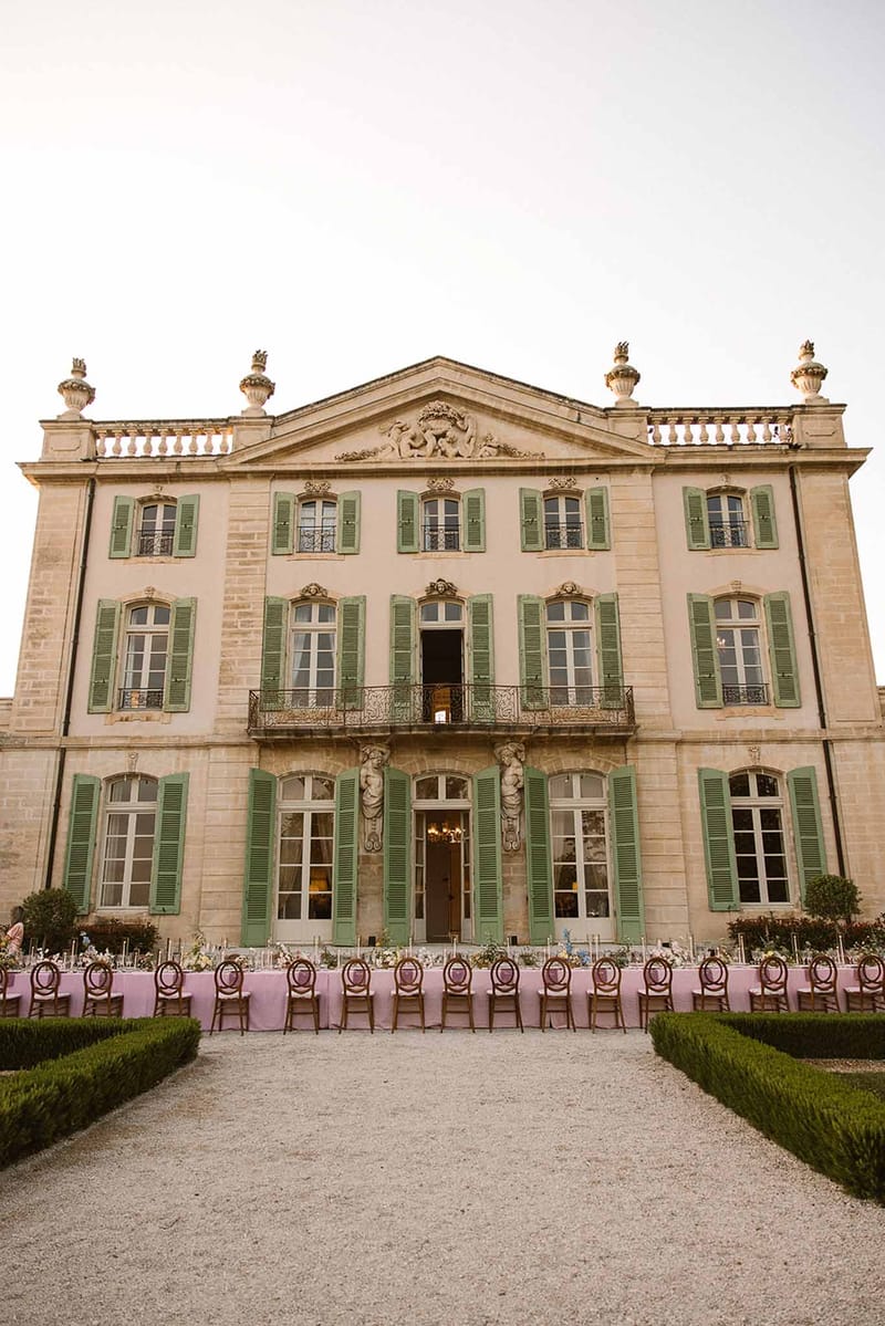 Long banquet table with blush linens and taper candles set on gravel forecourt in front of three-story stone chateau