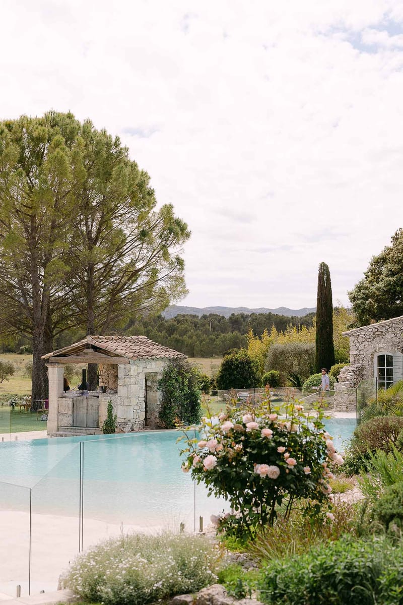 Provencal venue infinity pool with glass panels, stone poolhouse with terracotta roof, and blush pink rose bushes