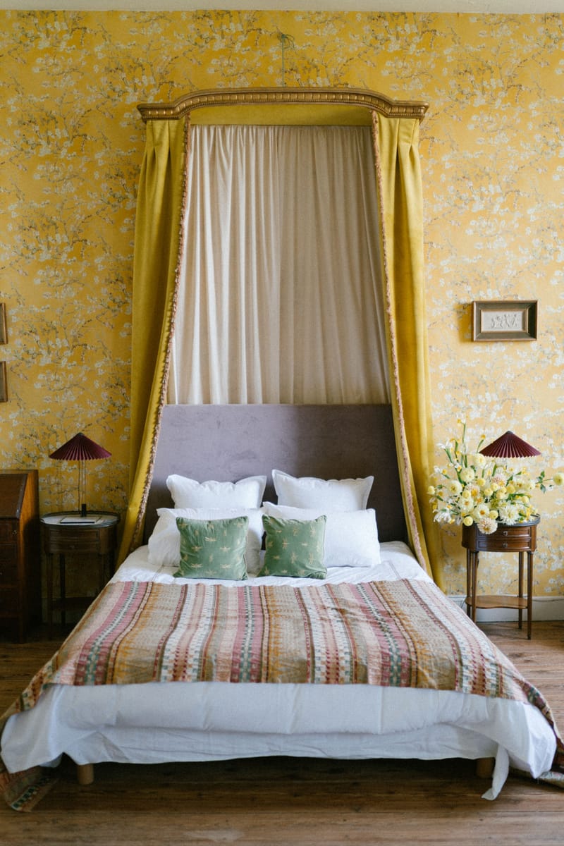 Chateau bedroom with yellow floral wallpaper, gilt canopy bed, sage cushions, and daffodil arrangement on nightstand