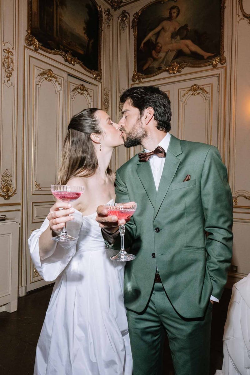 Bride and groom kissing while holding pink cocktails inside ornate French chateau with gold boiserie paneling