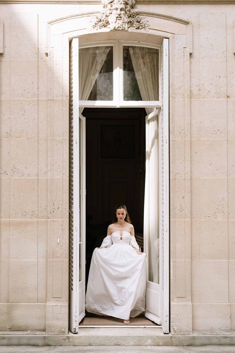 Bride in off-shoulder V-neck gown with puff sleeves standing in arched limestone doorway at chateau