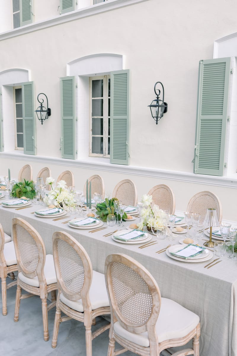 Grey linen table with white hydrangea runner, sage tapers, and cane-back chairs before green-shuttered manor