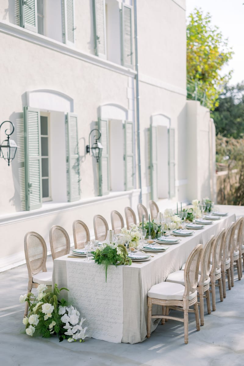 Long reception table with green taper candles, white roses, and cane-back chairs at white manor