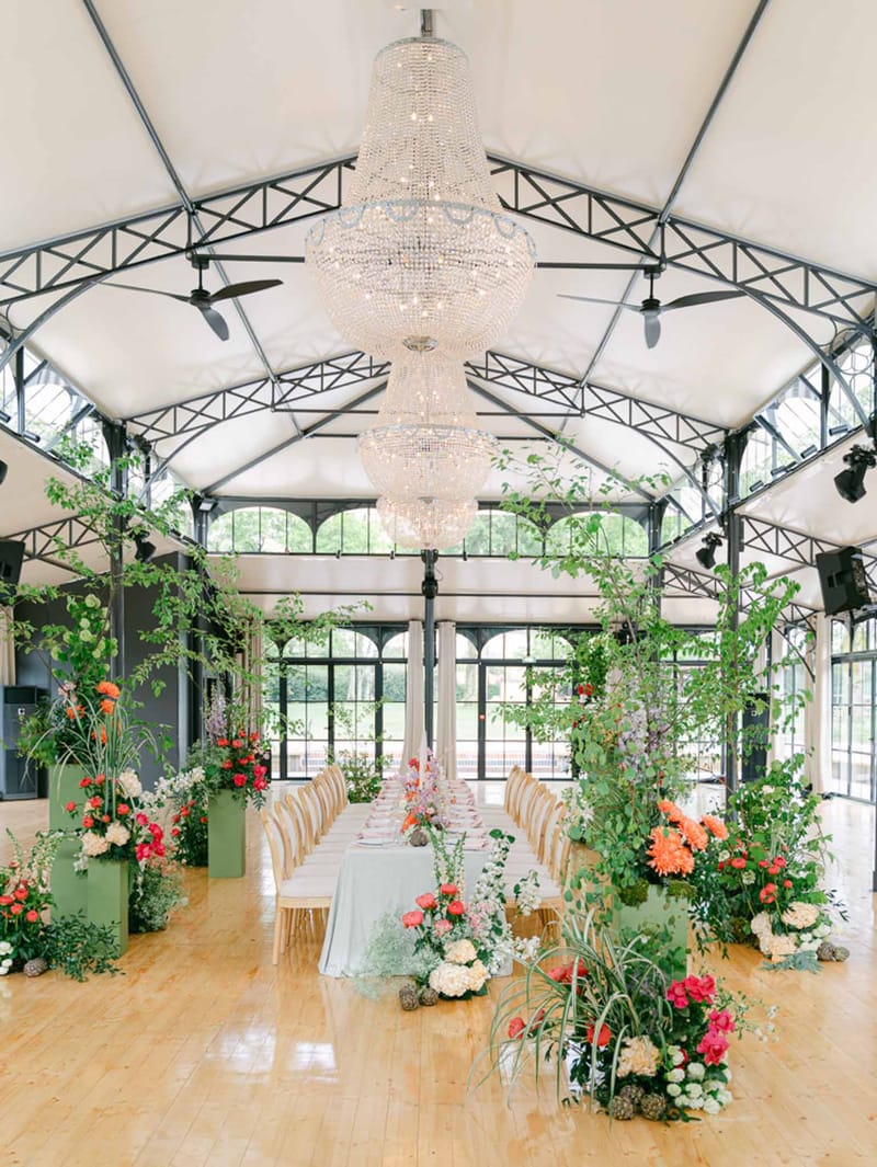 Greenhouse pavilion reception with long table, crystal chandeliers, sage planters, and coral and pink floral arrangements