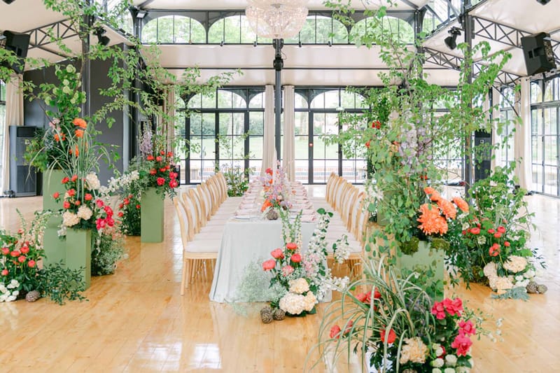 Orangerie reception with sage plinths holding coral dahlia and hot pink ranunculus arrangements and crystal chandelier