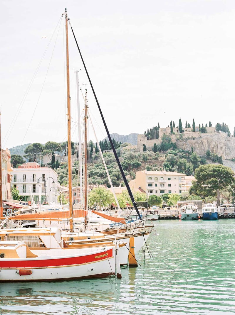 Mediterranean harbor with traditional sailboats moored in turquoise water, hillside castle and cypress trees behind