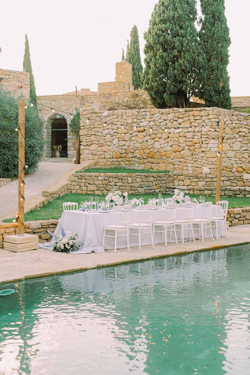 Long white table with hydrangea and blue floral cascade beside pool with globe lights at stone venue