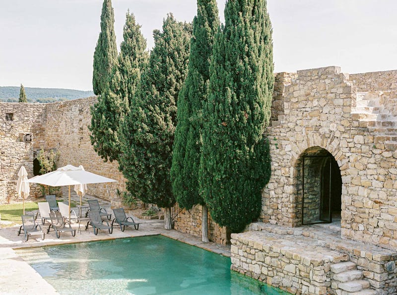 Limestone walled pool area with turquoise water grey loungers cream parasols cypress trees and stone archway with glass doors