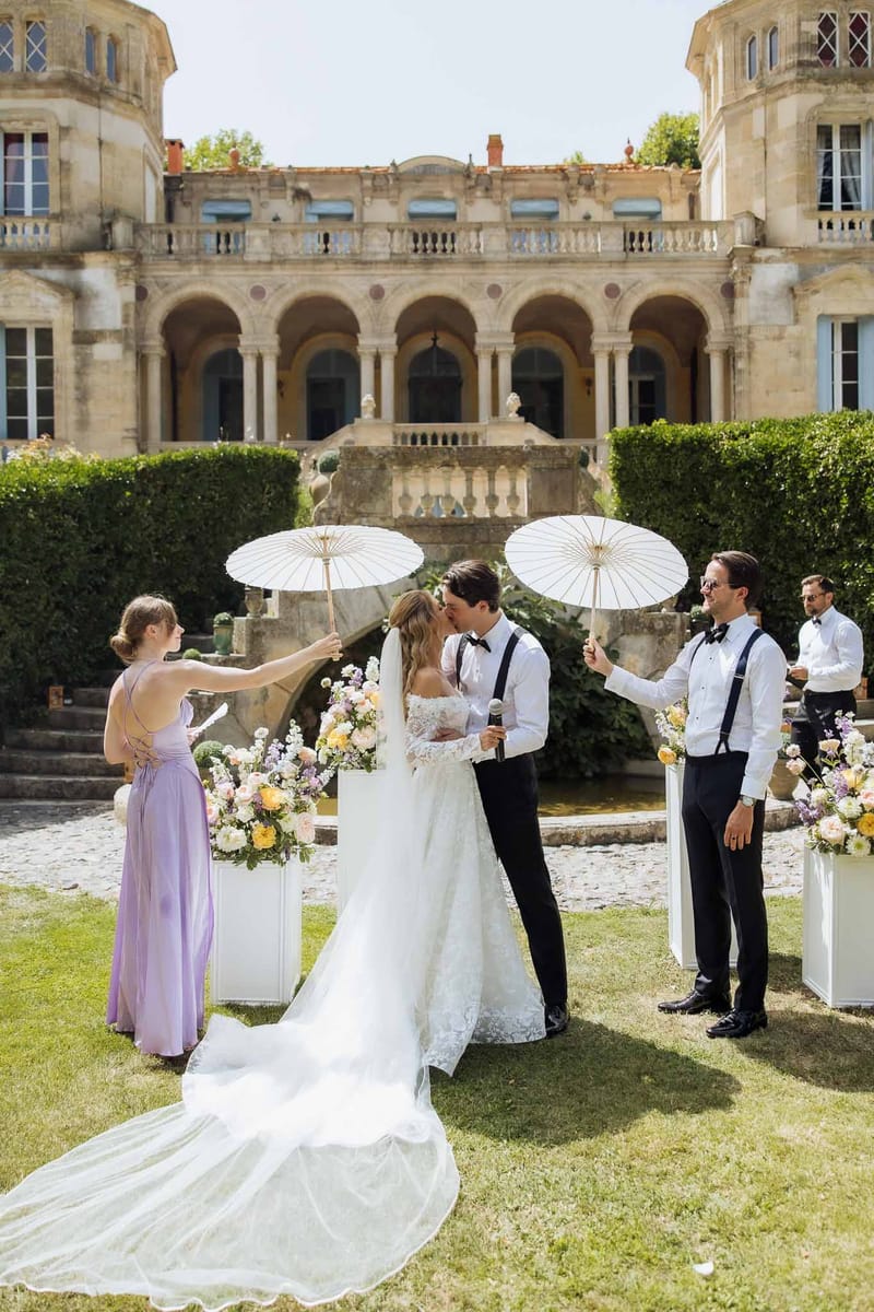 Bride and groom share first kiss on chateau lawn shaded by parasols with peach and lavender floral pedestals