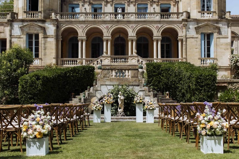 Empty ceremony setup with cross-back chairs and lavender ribbons facing chateau with floral pedestals
