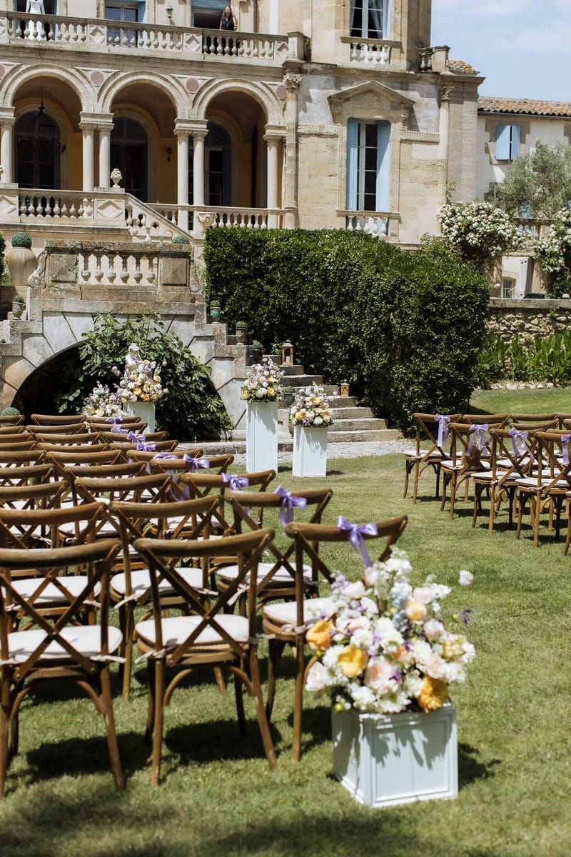 Ceremony setup with cross-back chairs, purple bows, and mixed floral pedestals at chateau