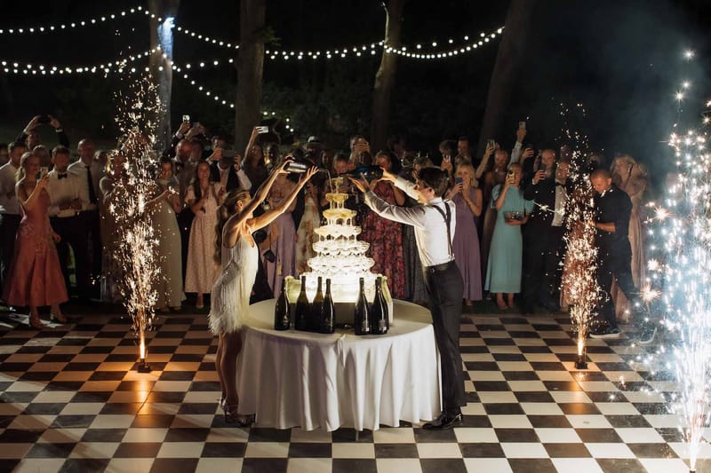 Couple pouring champagne tower on checkered dance floor with sparkler fountains and string lights at night