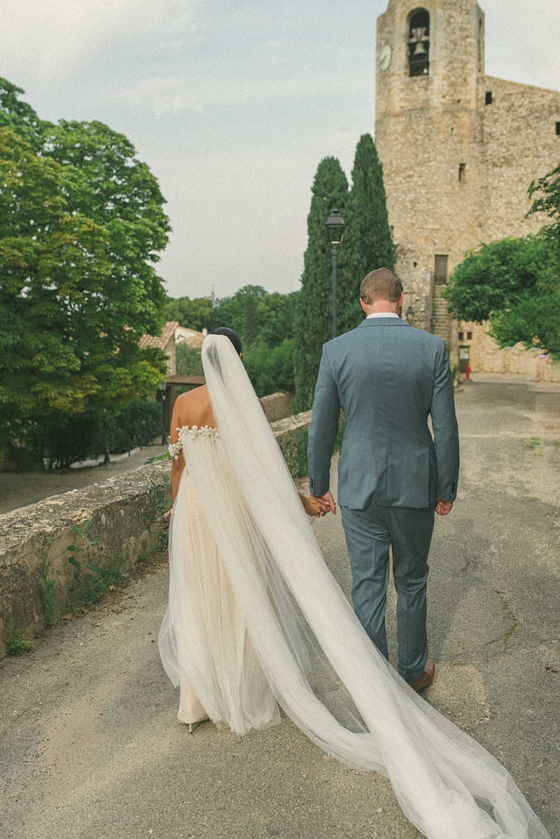 Couple walking hand-in-hand toward stone church, bride's cathedral veil trailing on cobblestone path