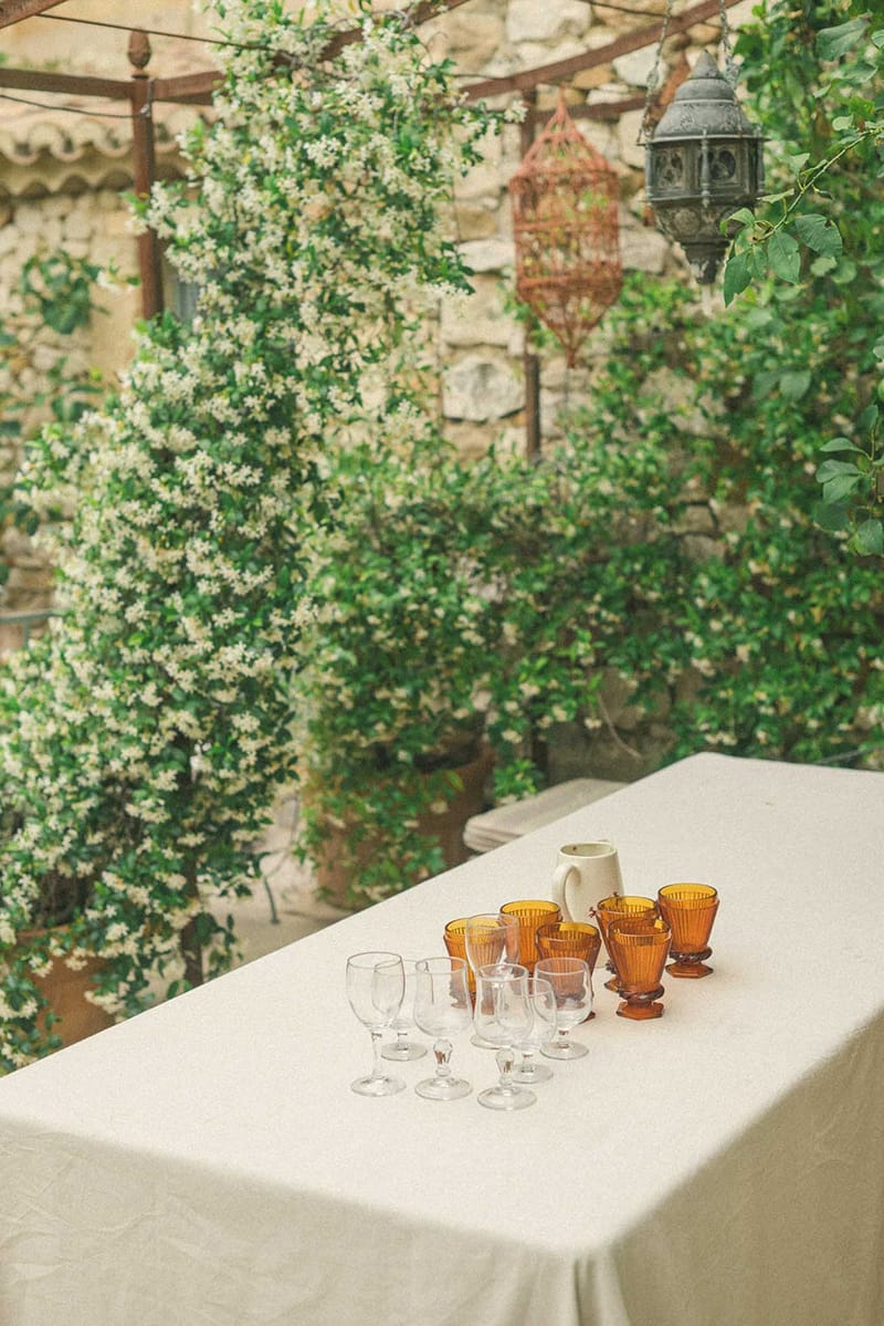 Outdoor bar table with amber ribbed goblets and clear stemware under hanging lanterns against vine-covered stone wall