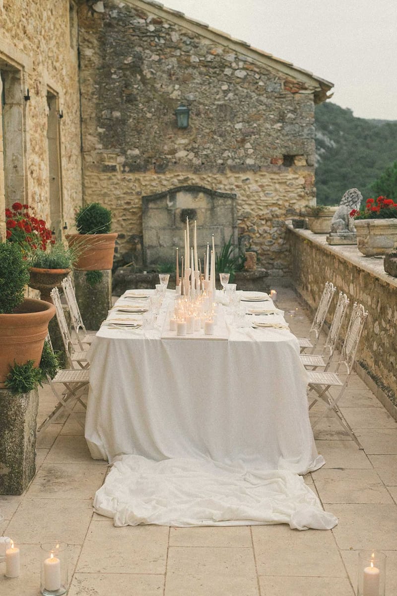 Candlelit banquet table with ivory tapers, gold-rimmed plates, and white folding chairs on stone terrace