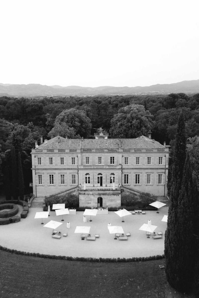 Black and white aerial classical manor with white market umbrellas on forecourt clipped hedgerows and cypress trees