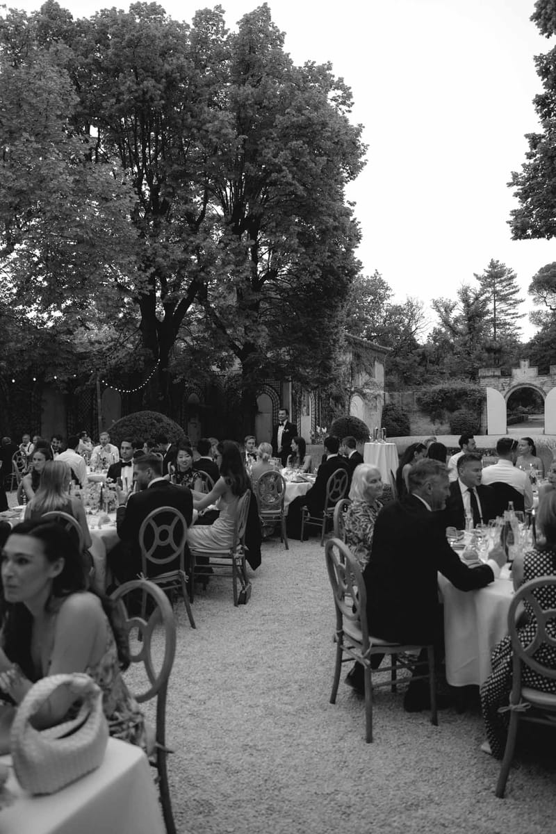 Black and white wide shot of outdoor reception dinner with guests at round tables under string lights