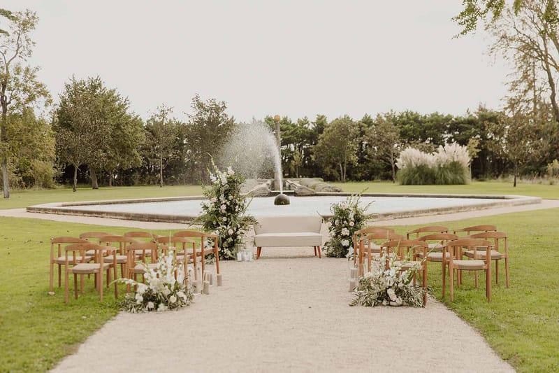 Ceremony aisle with wood chairs white and blush florals leading to cream sofa altar before stone fountain