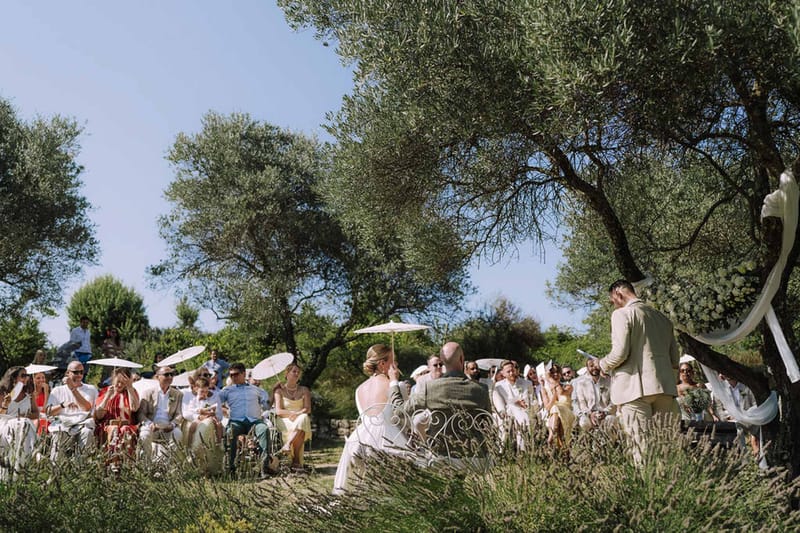 Ceremony in olive grove with wine barrel podium, rustic arch with coral flowers, and white parasol guests