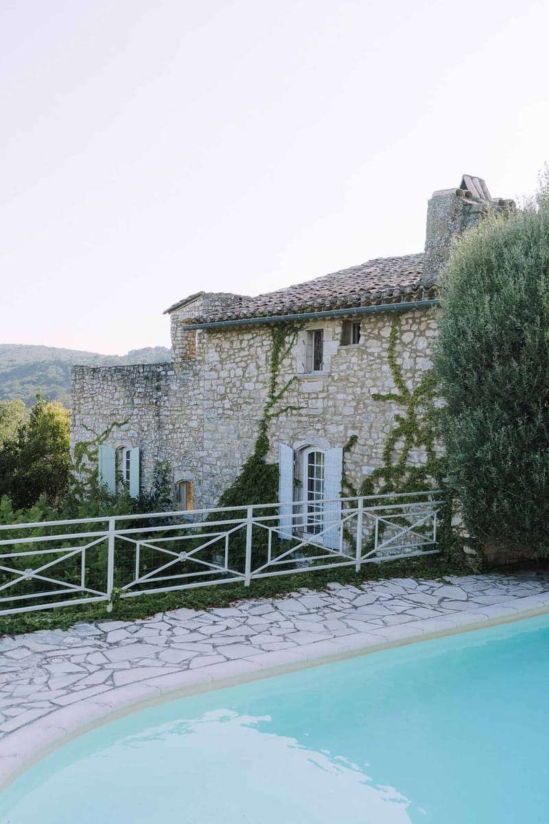 Provencal stone farmhouse with terracotta roof, blue-grey shutters, and climbing ivy beside turquoise pool on stone terrace