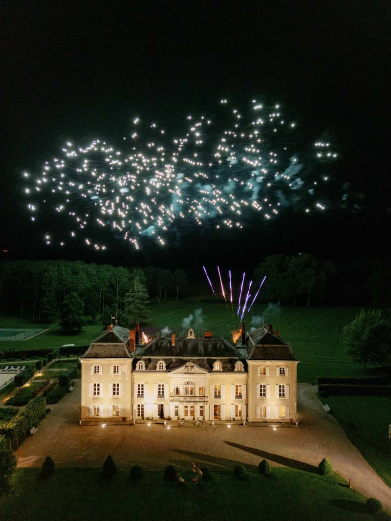 Aerial night view of chateau with rooftop fireworks, formal parterre garden, and illuminated facade