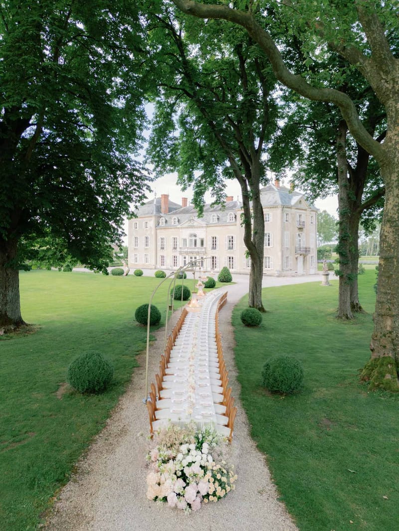 Long feasting table with blush rose runner and brass arch on gravel path leading to cream stone chateau