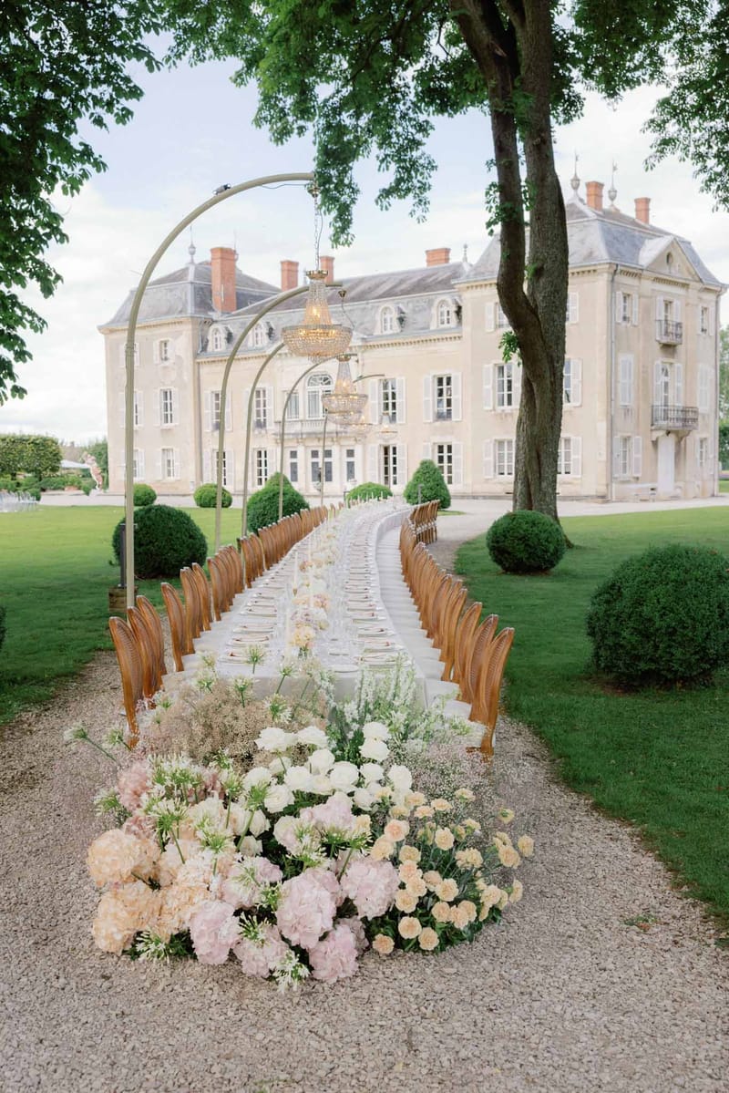 Long reception table with brass arches and chandeliers leading toward French chateau, pastel floral runner