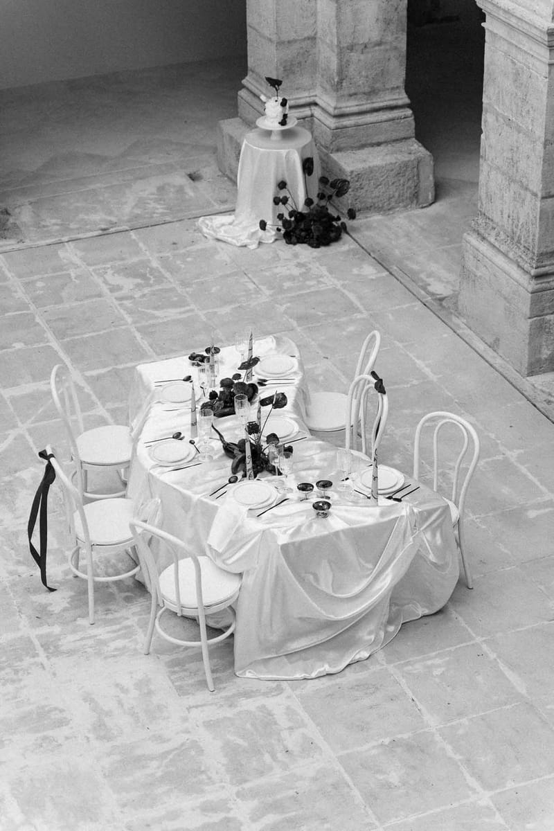 Black-and-white styled reception table with dark floral centerpiece, taper candles, and single-tier cake in stone courtyard