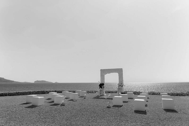 Black and white image of minimalist outdoor ceremony setup with white cube seating on terrace overlooking the sea