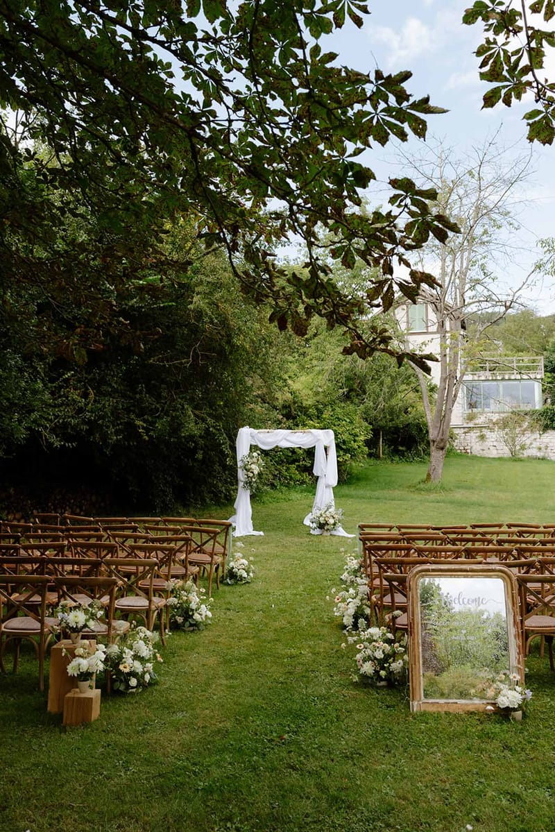Outdoor ceremony setup with wooden crossback chairs, white floral aisle decor, draped wooden arch, and gold welcome mirror