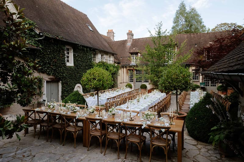 Banquet tables with white linens and brass candlesticks in ivy-covered French manor courtyard with topiary