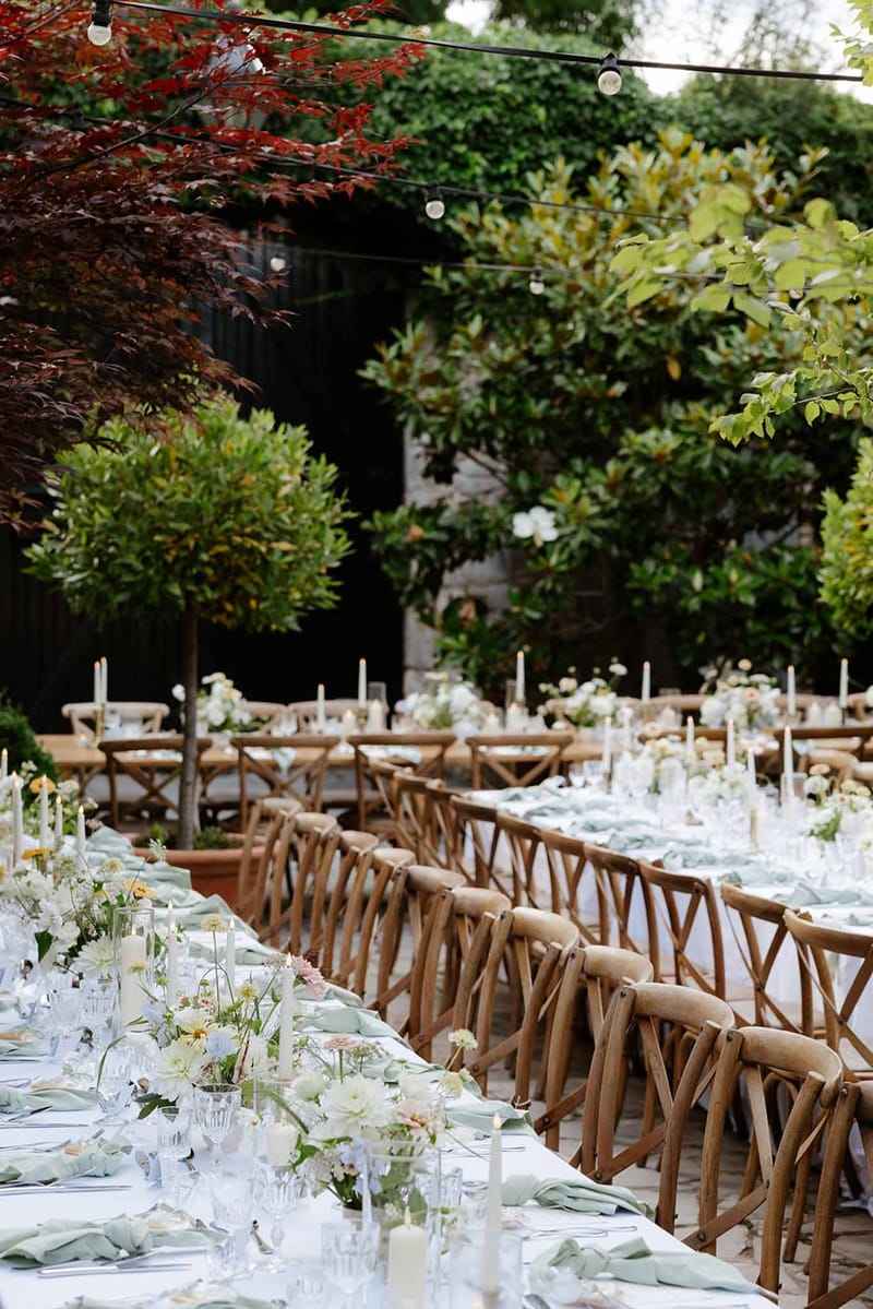 Outdoor reception with sage napkins, wildflower arrangements, and Edison lights in garden courtyard