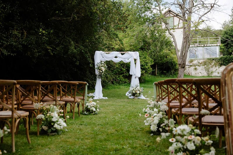 Garden ceremony aisle with fabric-draped wooden arch, cross-back chairs, and white dahlia arrangements on lawn