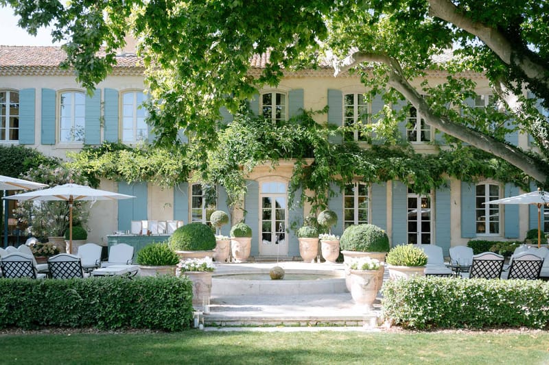 Provencal mas with pale stone walls, blue shutters, topiary pots, and terrace set for cocktail hour