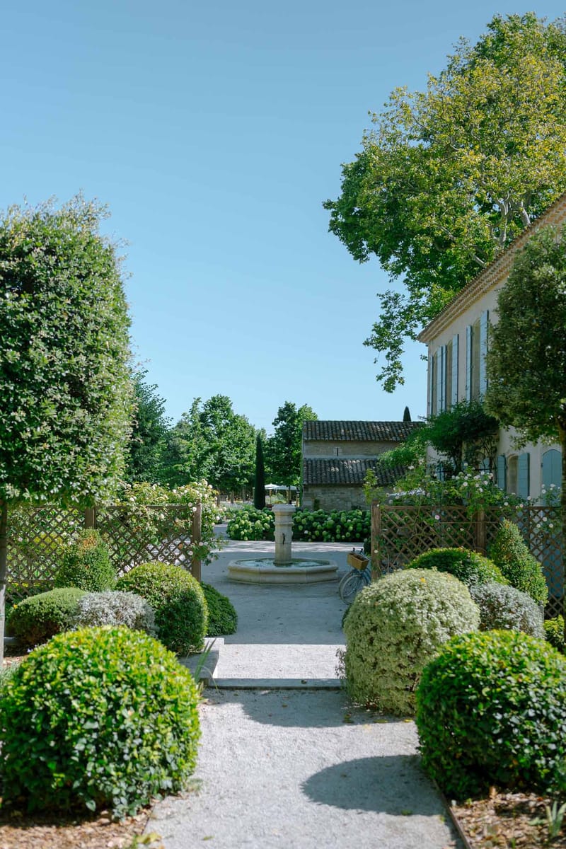 Provencal garden with box ball topiaries, stone pedestal fountain, and two-storey mas with blue shutters