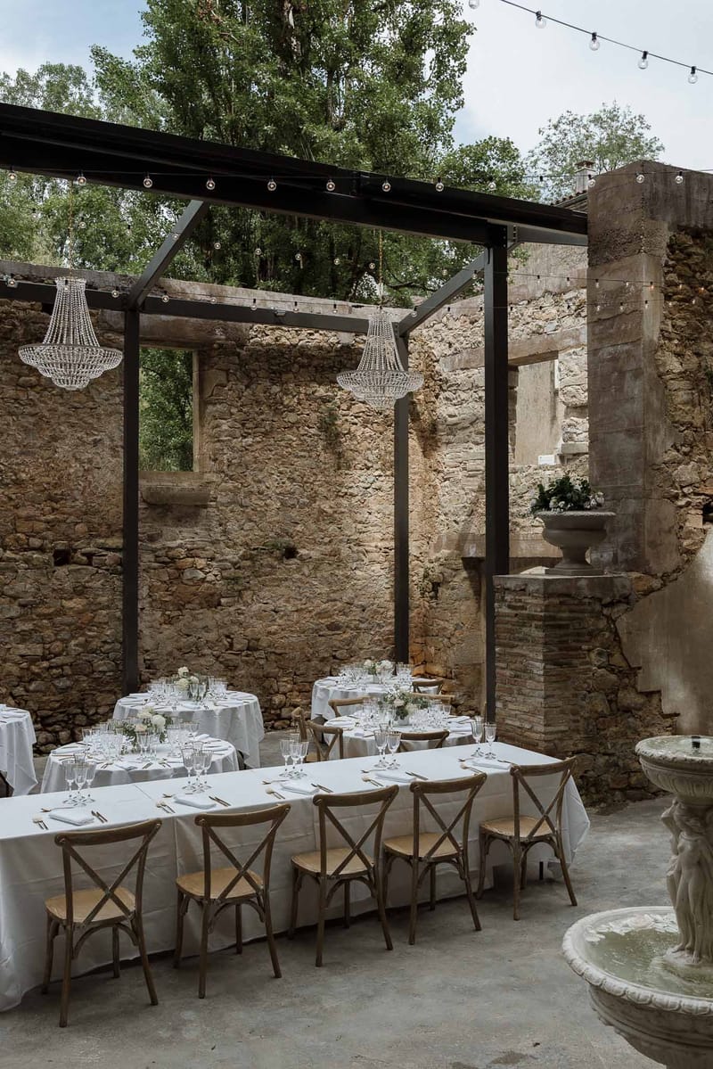 Wedding reception in ruined stone courtyard with black pergola, Edison bulb lights, crystal chandeliers, and white-dressed...