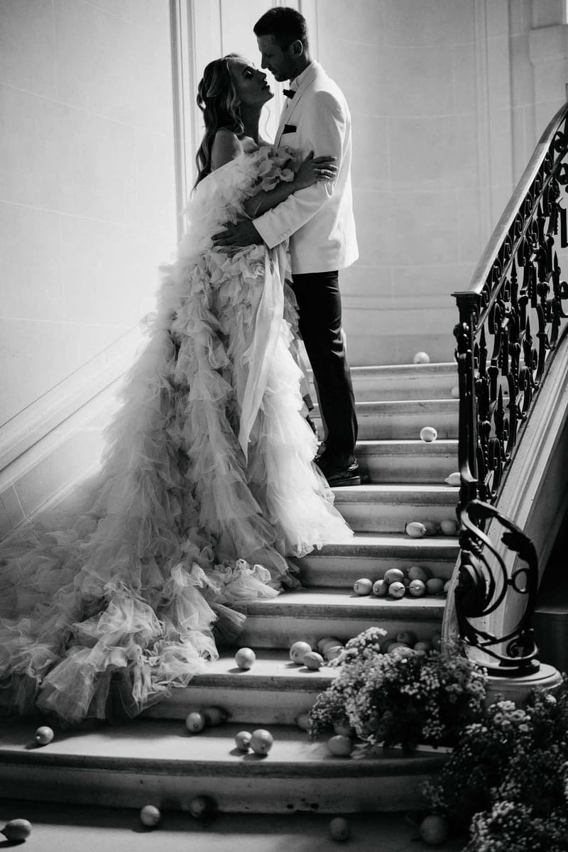 Black and white portrait of bride and groom embracing on grand chateau staircase with ruffled tulle train cascading down s...