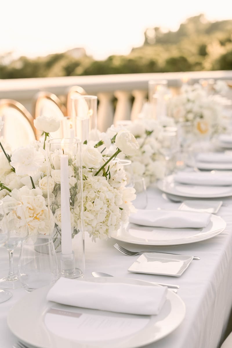 All-white tablescape with hydrangea and rose runner, taper candles in hurricane holders, and gold chairs on terrace