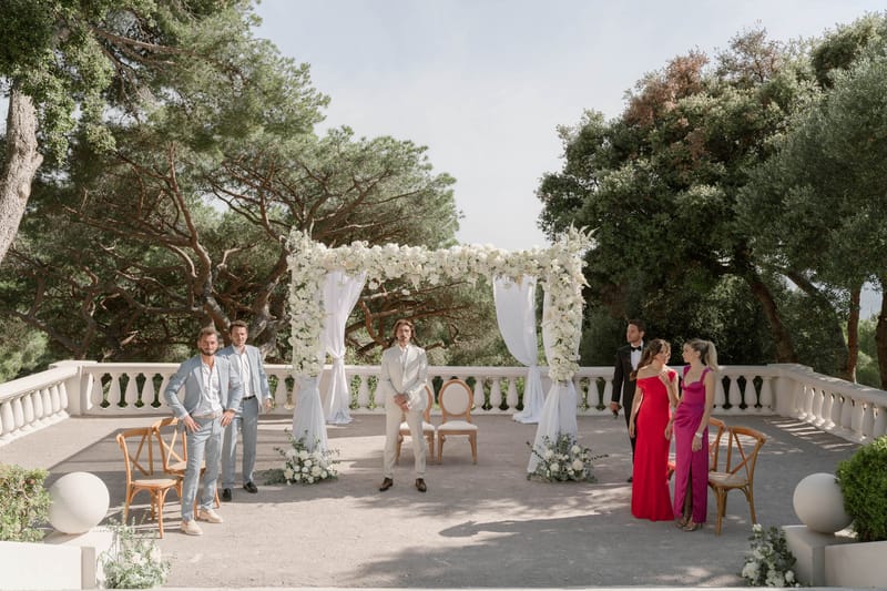 Ceremony setup on balustraded terrace with white rose and orchid draped arch and seated groom in cream suit