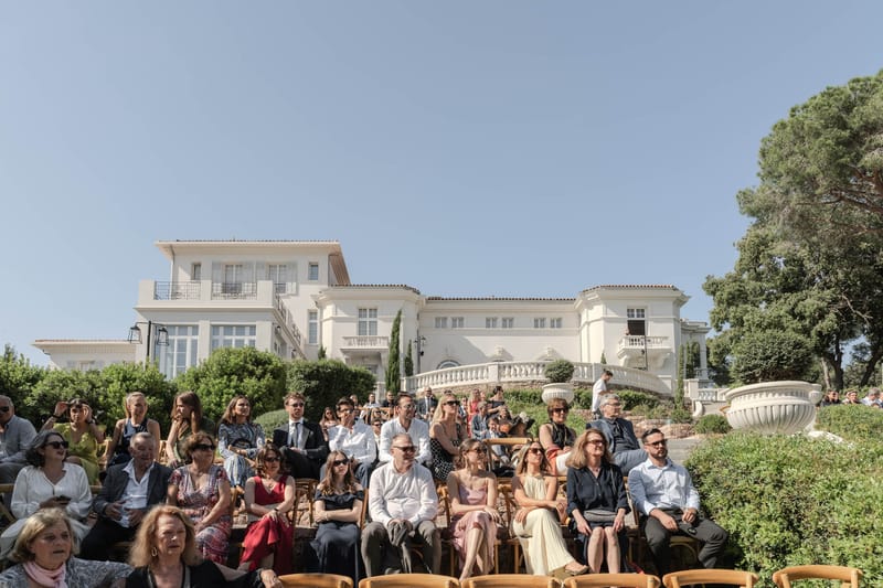 Forty guests seated for outdoor ceremony before white rendered villa with cypress trees and balustrades