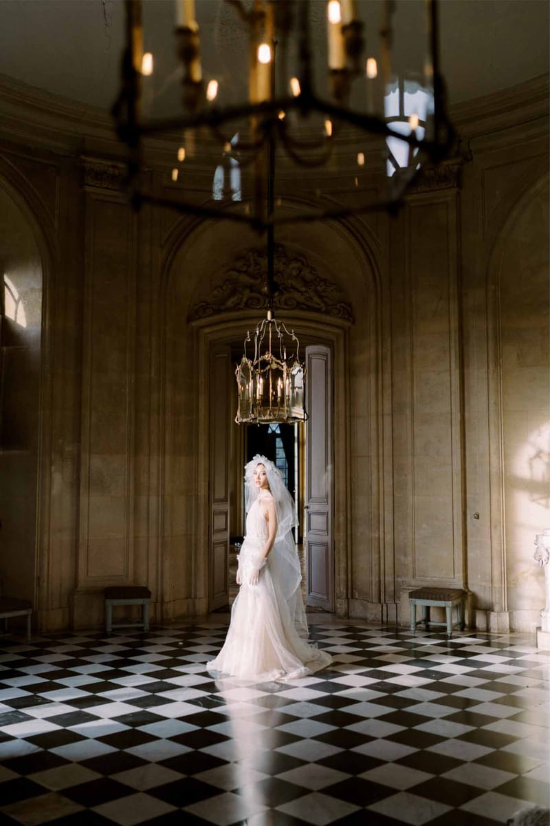 Bride in blush tulle gown and Juliet cap veil on marble diamond floor beneath brass chandeliers
