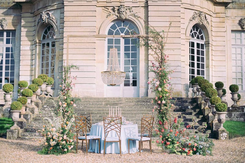 Styled sweetheart table under gold chandelier arch with coral and peach floral columns before chateau