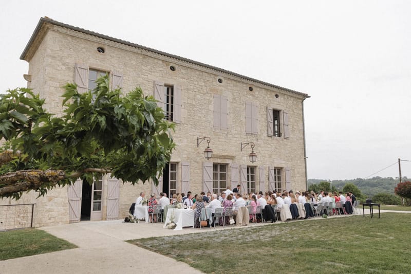 Fifty guests at long table before Provencal stone farmhouse with taupe shutters and wrought-iron lanterns