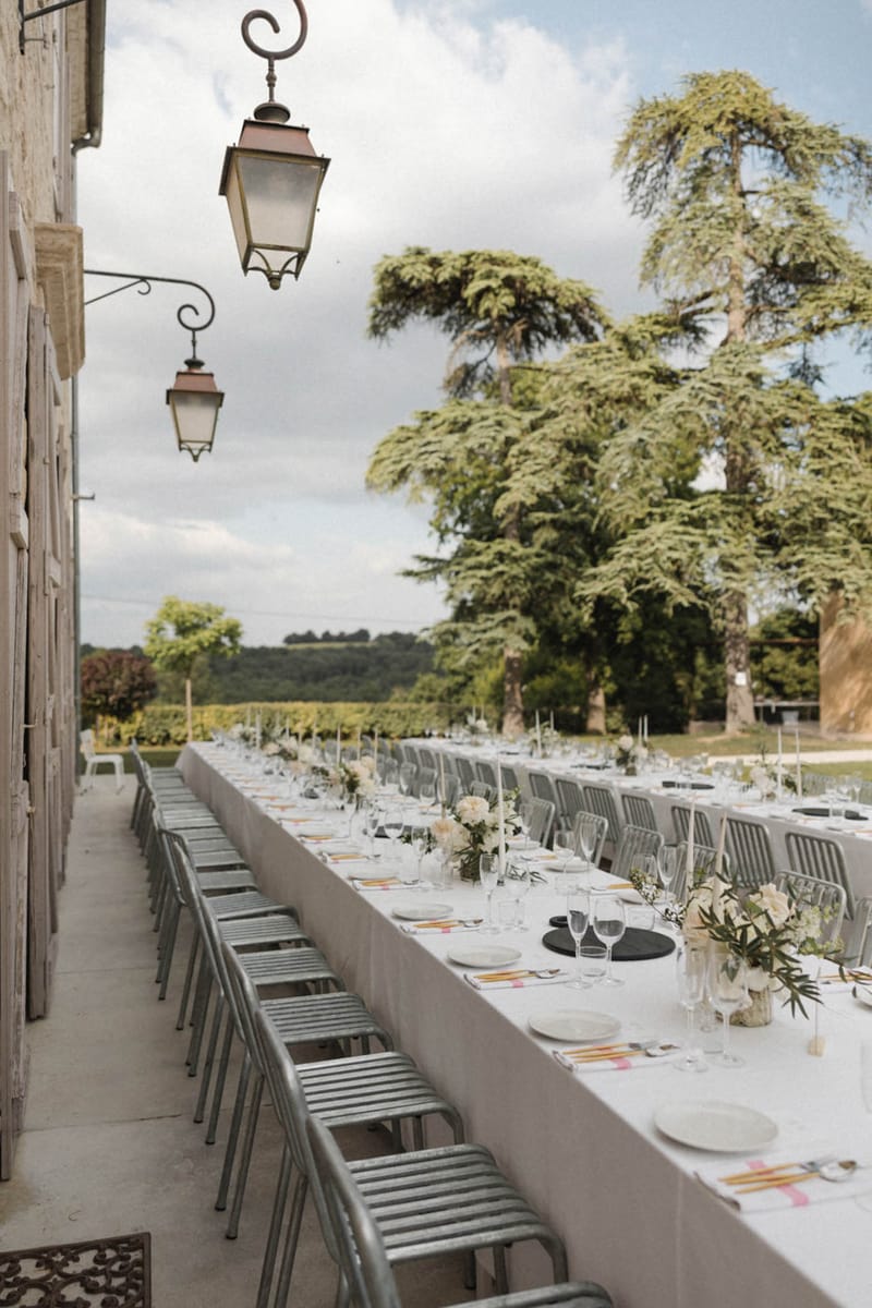 Two long banquet tables on chateau terrace with sage chairs, white linens, and low floral centerpieces