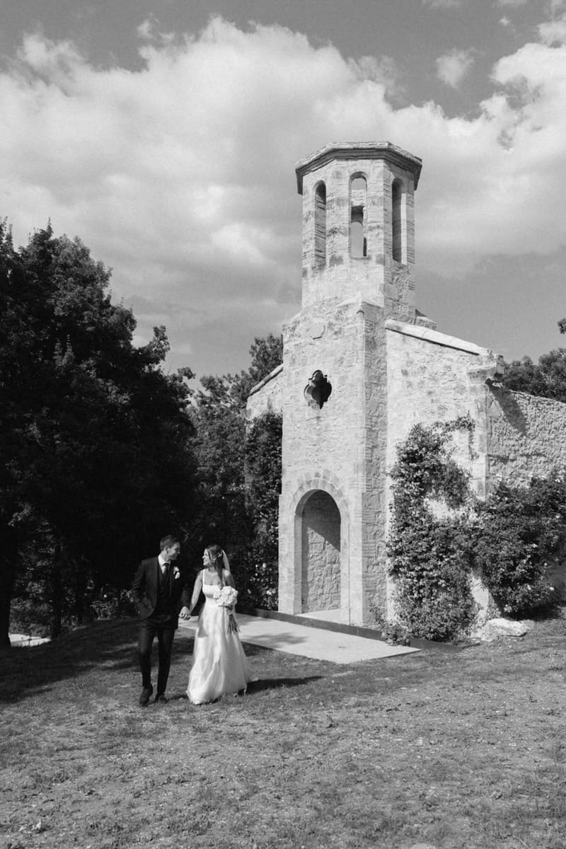 Black-and-white portrait of bride and groom walking hand in hand past a Romanesque stone chapel with octagonal bell tower